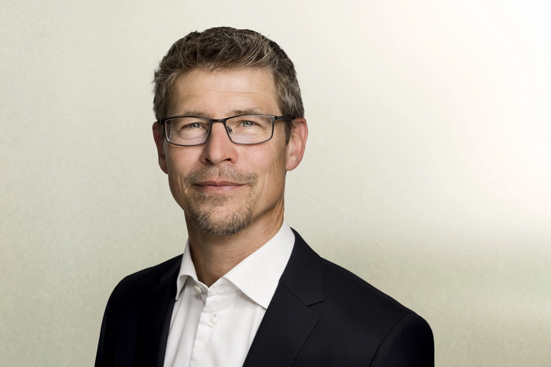 Mężczyzna w okularach, uśmiechający się, ubrany w garnitur na tle w jasnym kolorze.