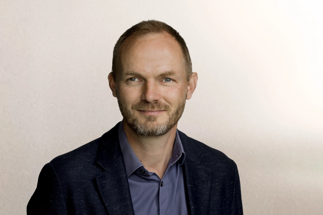 Mężczyzna z zarostem, uśmiechający się, w eleganckim ubraniu, na neutralnym tle.