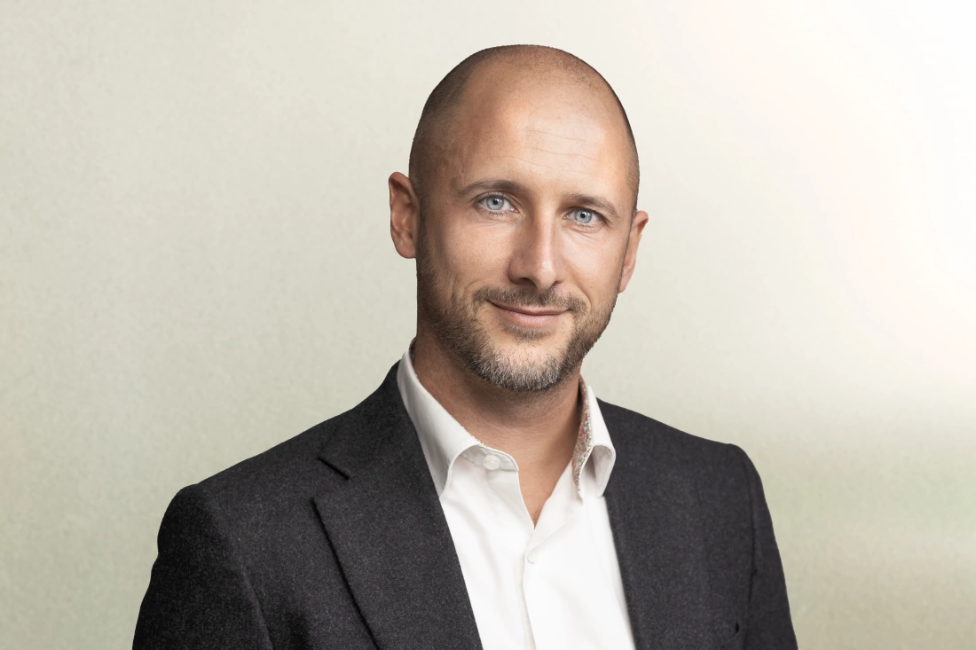 Mężczyzna w garniturze z uśmiechem, stojący na tle o stonowanej barwie.