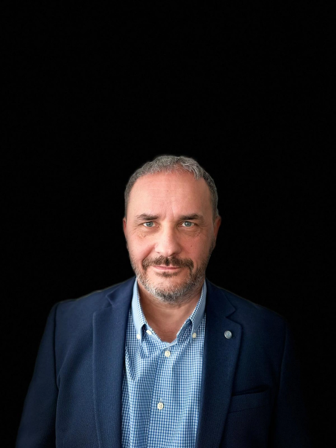 Mężczyzna o siwych włosach, ubrany w granatową marynarkę i koszulę w kratę, na tle czarnego.