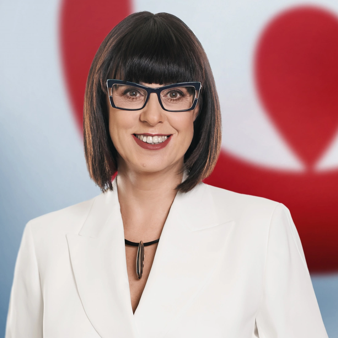 Kobieta z krótkimi włosami, w okularach i białej marynarce, uśmiechająca się na tle kolorowego tła.