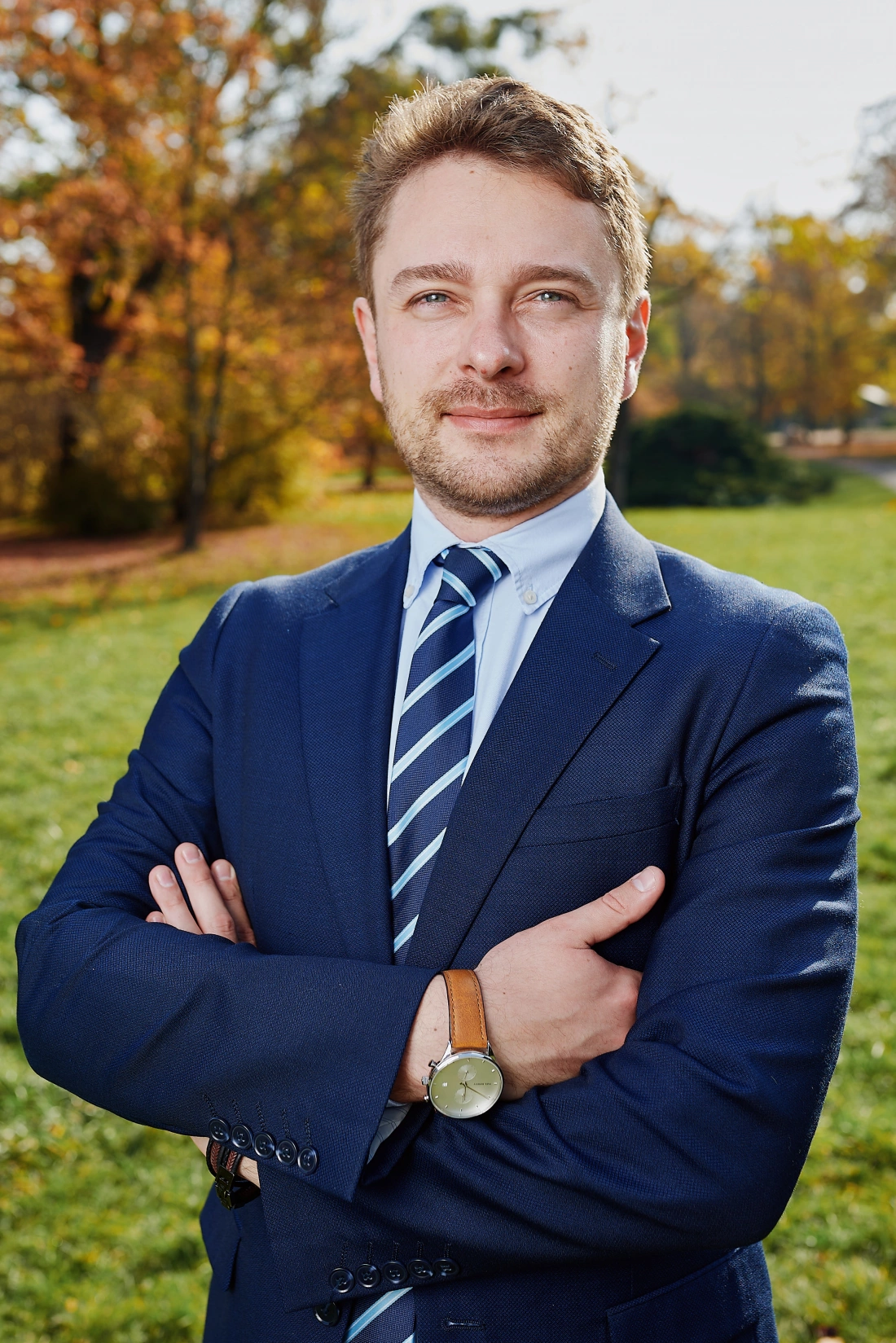 Mężczyzna w garniturze z zegarkiem, stojący z założonymi rękami na tle parku w słoneczny dzień.