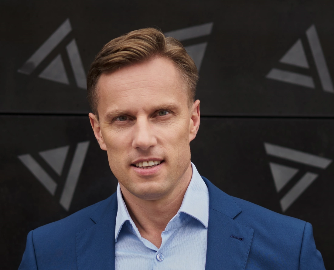 Mężczyzna w niebieskim garniturze uśmiecha się na tle abstrakcyjnego, ciemnego wzoru.