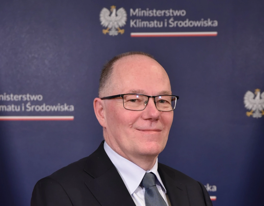 Mężczyzna w okularach, uśmiechający się przed tłem z logo Ministerstwa Klimatu i Środowiska.