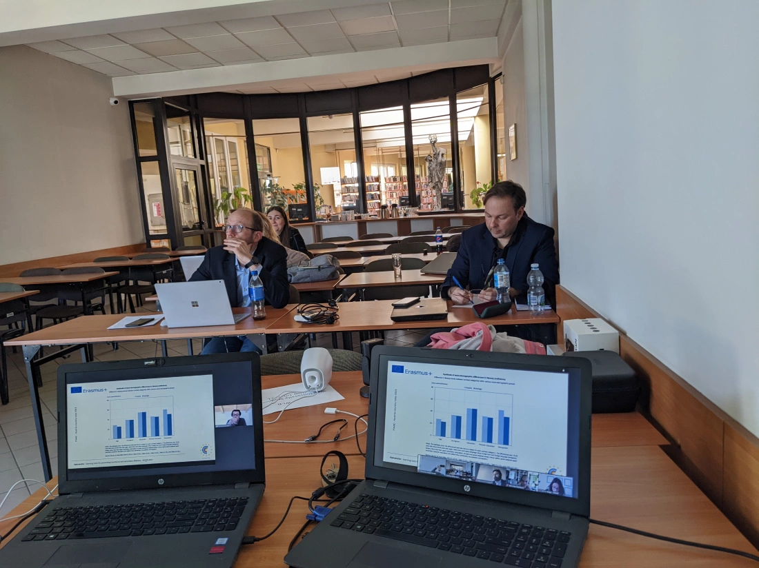 Spotkanie w sali z dwoma osobami przy stołach, na laptopach wykresy i prezentacja, widoczna biblioteka w tle.