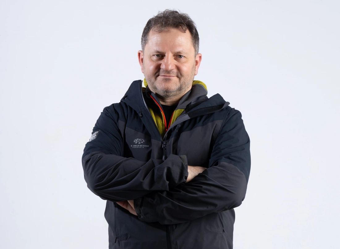 Mężczyzna w czarnej kurtce z założonymi rękami, uśmiechający się do aparatu na tle białej ściany.