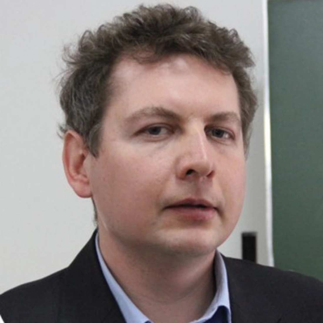 Mężczyzna z krótkimi, falującymi włosami, w ciemnym garniturze, spogląda w obiektyw.