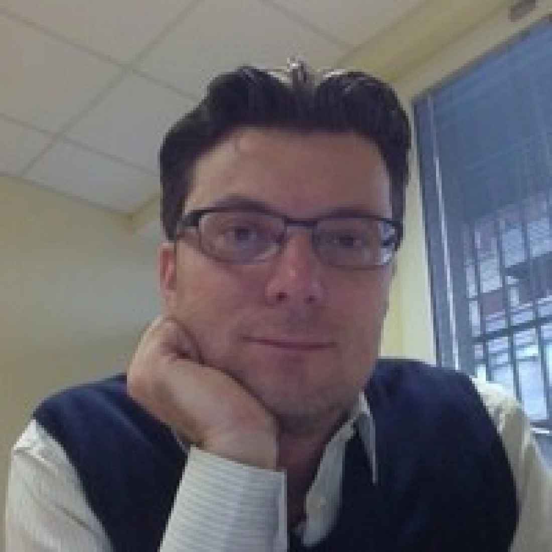 Mężczyzna w okularach, z ręką podłożoną pod brodę, siedzi w biurze.