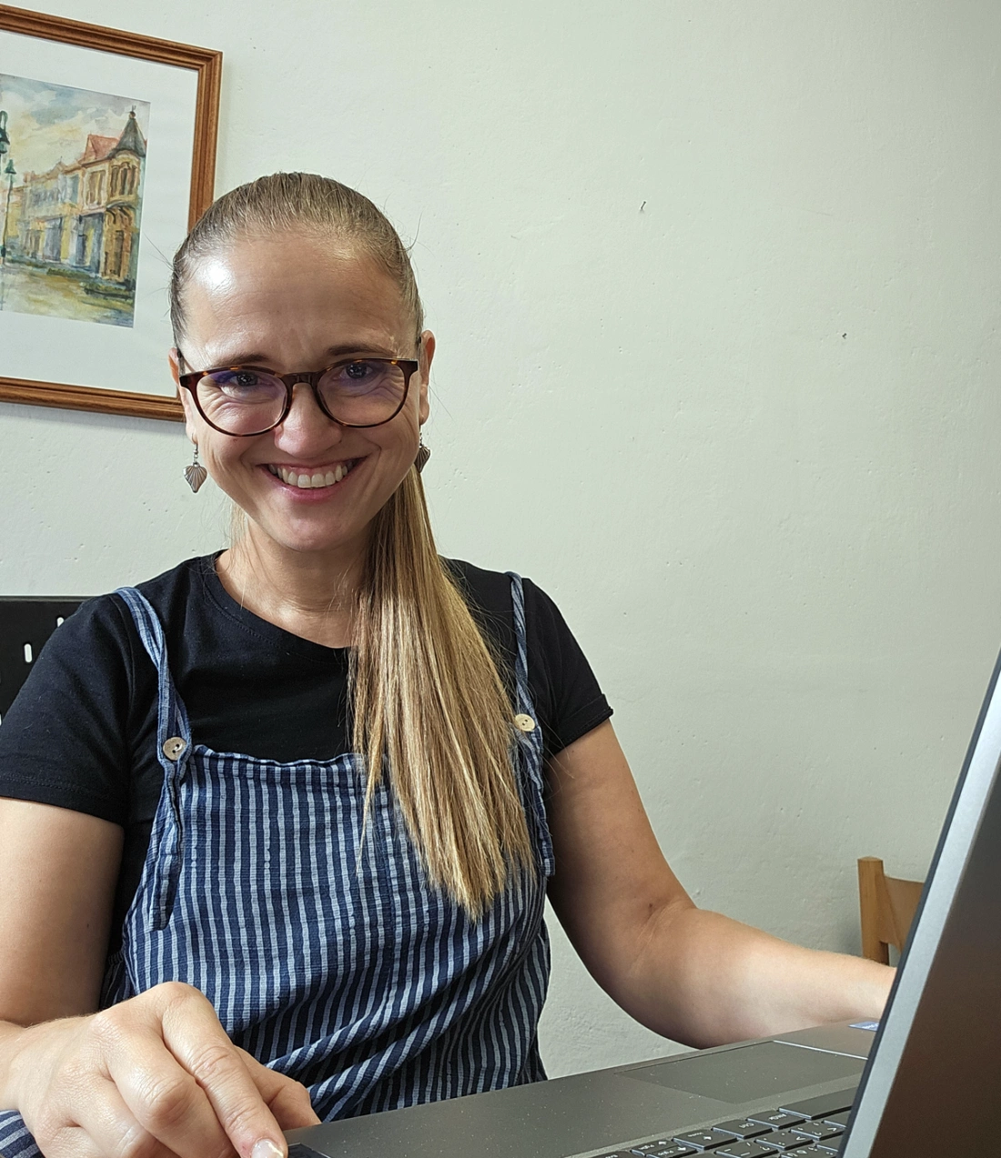 Uśmiechnięta kobieta siedzi przy biurku z laptopem, w tle widoczny jest obrazek.