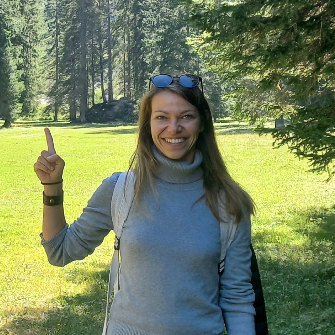 Kobieta w szarej bluzie stoi na tle lasu, wskazując na coś z uśmiechem.