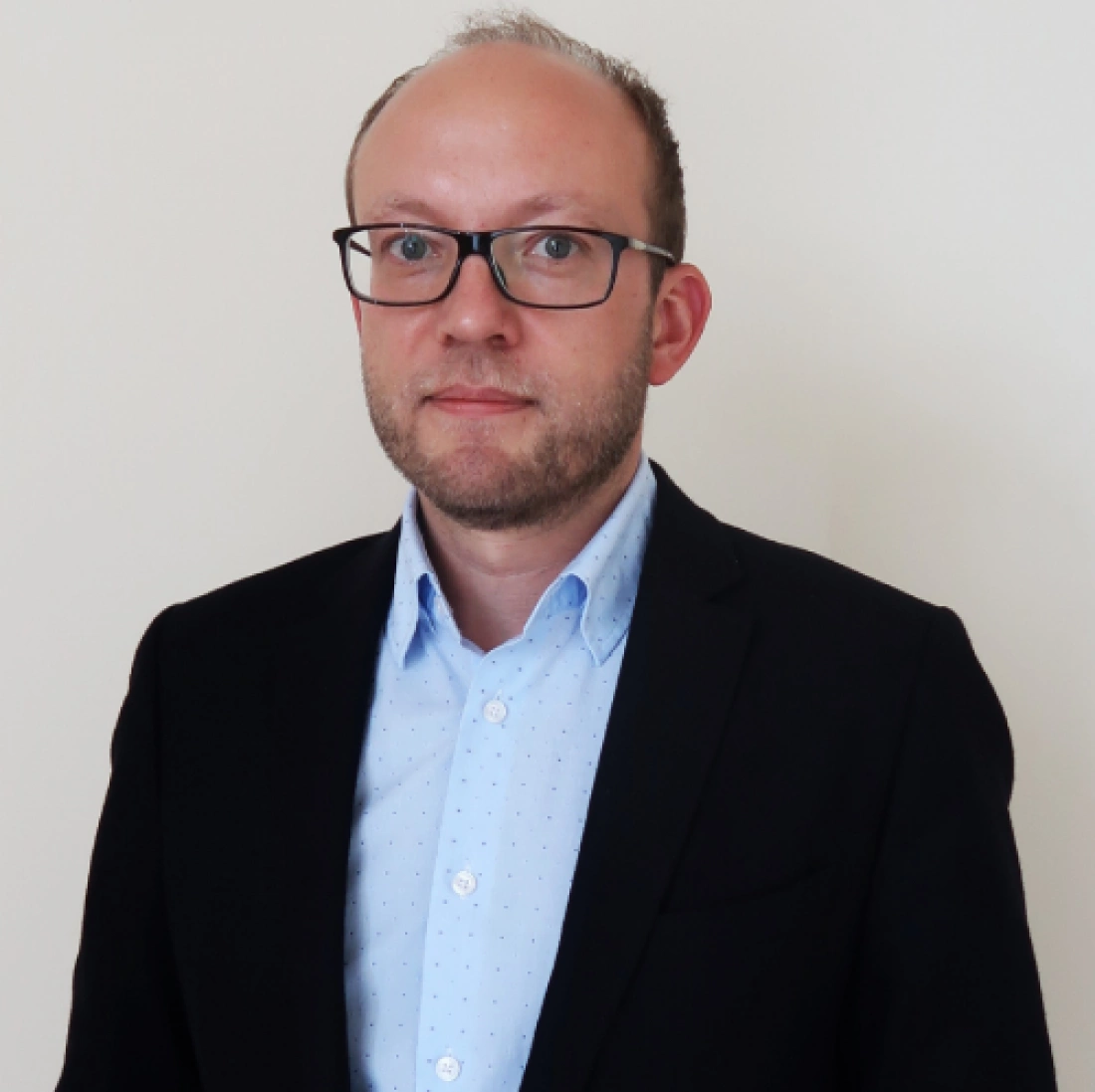 Mężczyzna w okularach, ubrany w marynarkę i koszulę, z neutralnym tłem.