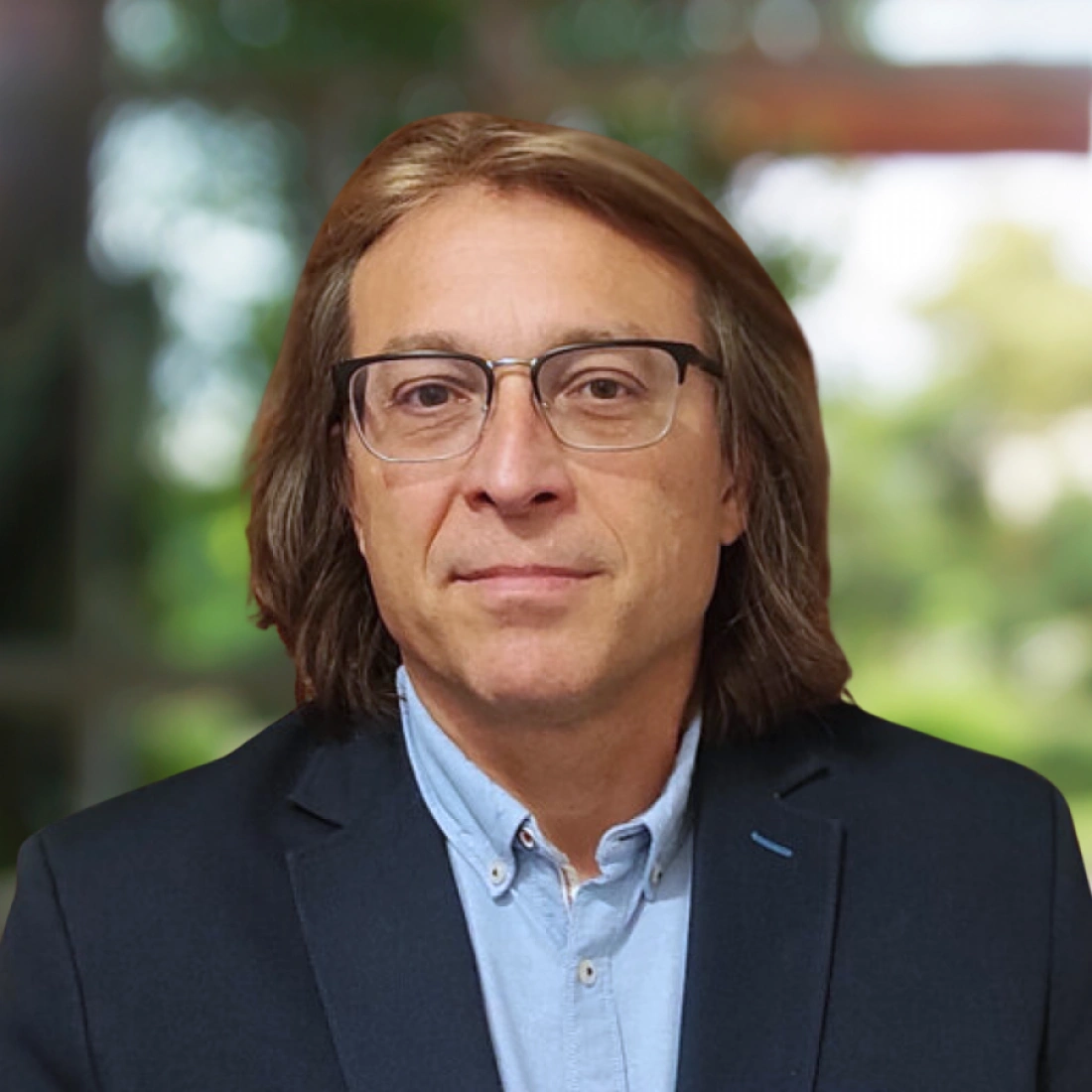 Mężczyzna w okularach, ubrany w ciemny żakiet i jasną koszulę, stoi na tle rozmytego krajobrazu.