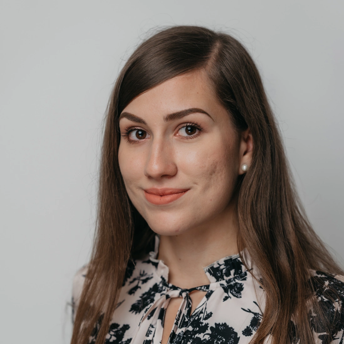 Portret młodej kobiety z długimi włosami, w kwiatującej bluzce, z delikatnym uśmiechem.