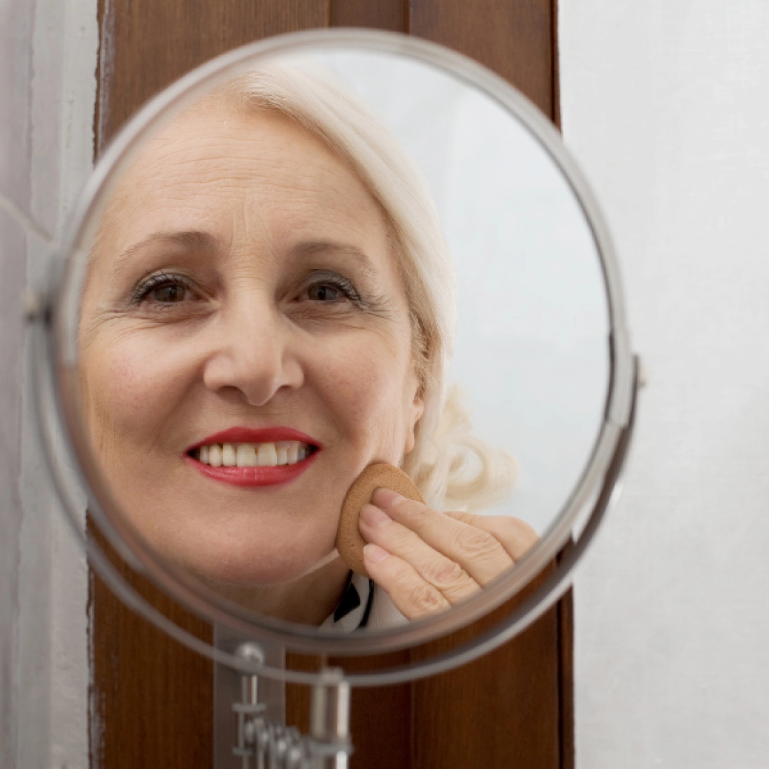 Kobieta z siwymi włosami uśmiecha się, aplikując kosmetyk w lustrze.