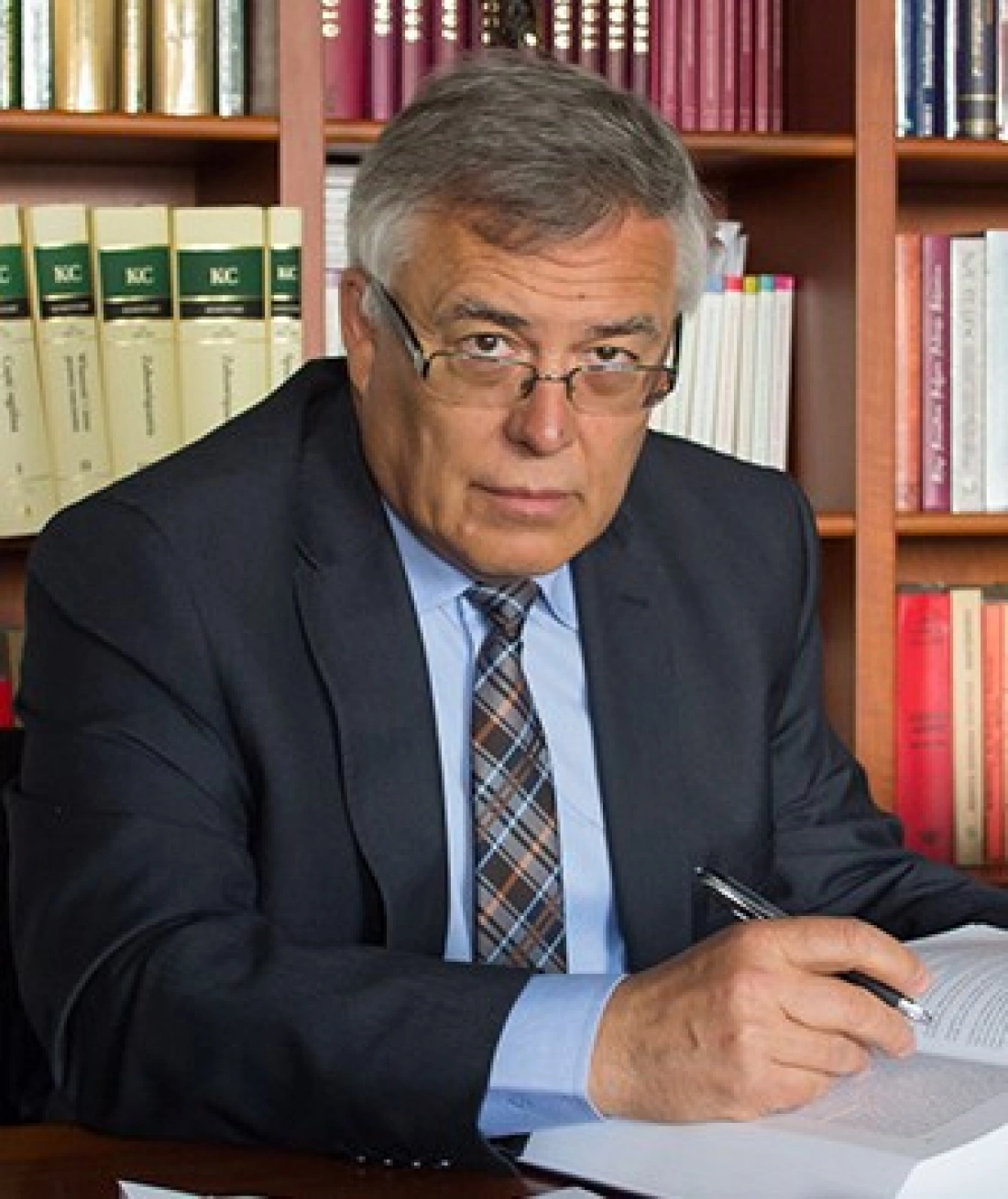 Mężczyzna w garniturze, siedzący przy biurku z dokumentami, pisze ołówkiem. W tle widoczne książki.