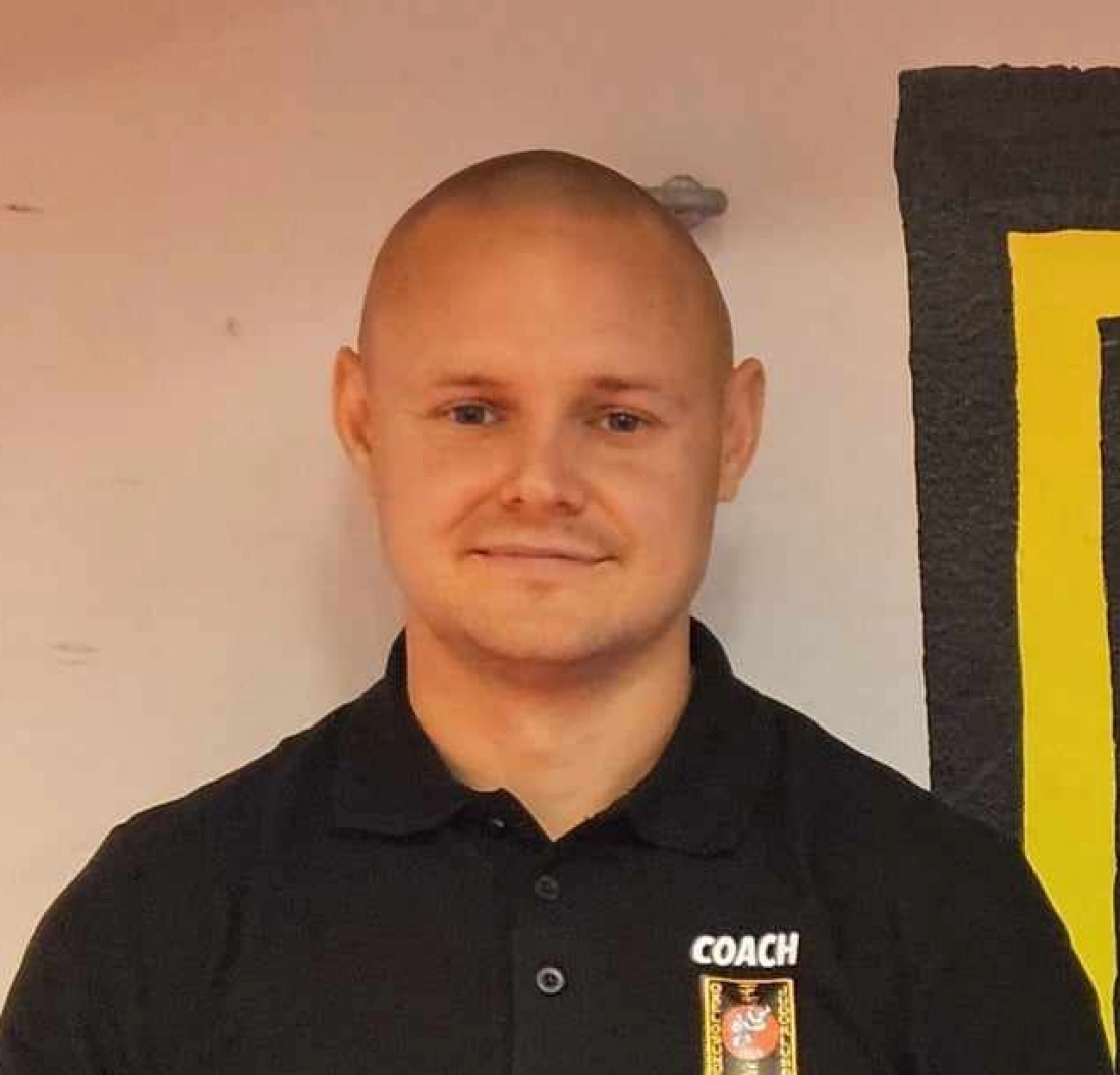 Mężczyzna z łysą głową, ubrany w czarną koszulę z napisem "COACH", stoi na tle kolorowej ściany.