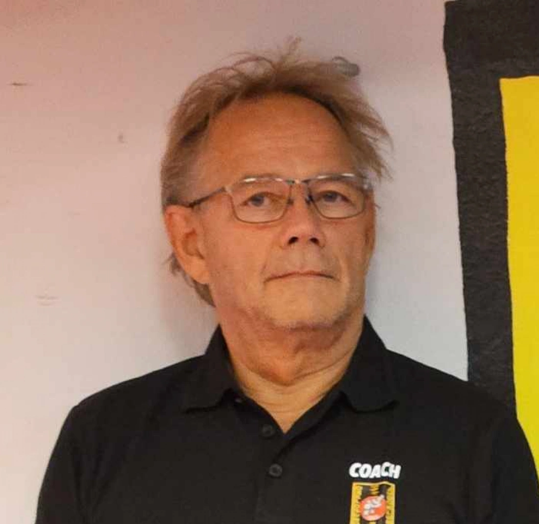 Mężczyzna w okularach z siwymi włosami, ubrany w czarną koszulkę z napisem "COACH", stoi w pomieszczeniu.