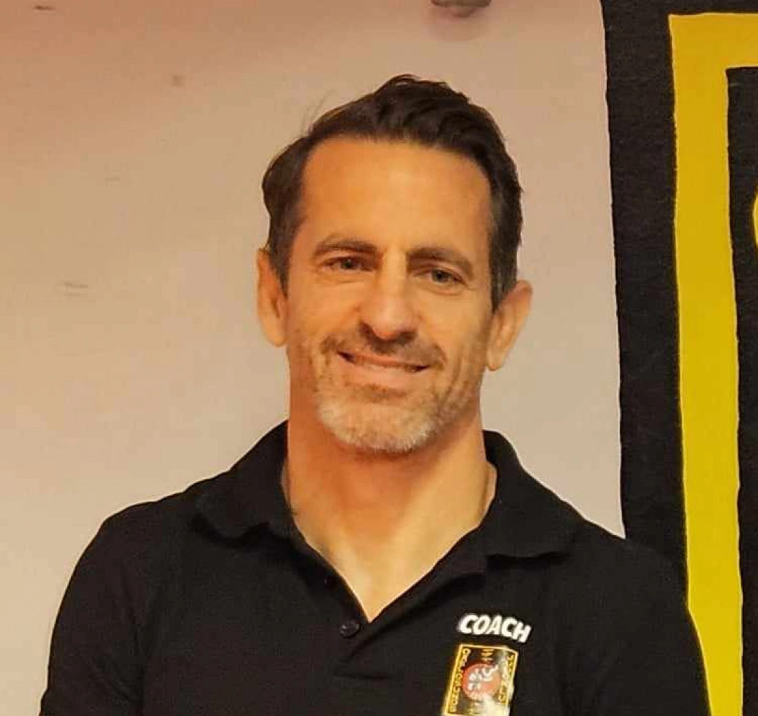 Mężczyzna w czarnej koszulce z napisem "COACH", uśmiecha się na tle kolorowej ściany.