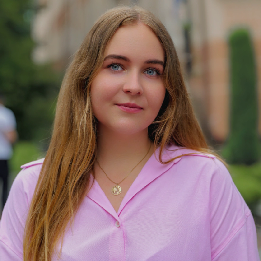 Młoda kobieta z długimi włosami, ubraną w różową bluzkę, stoi na tle zielonego krajobrazu.