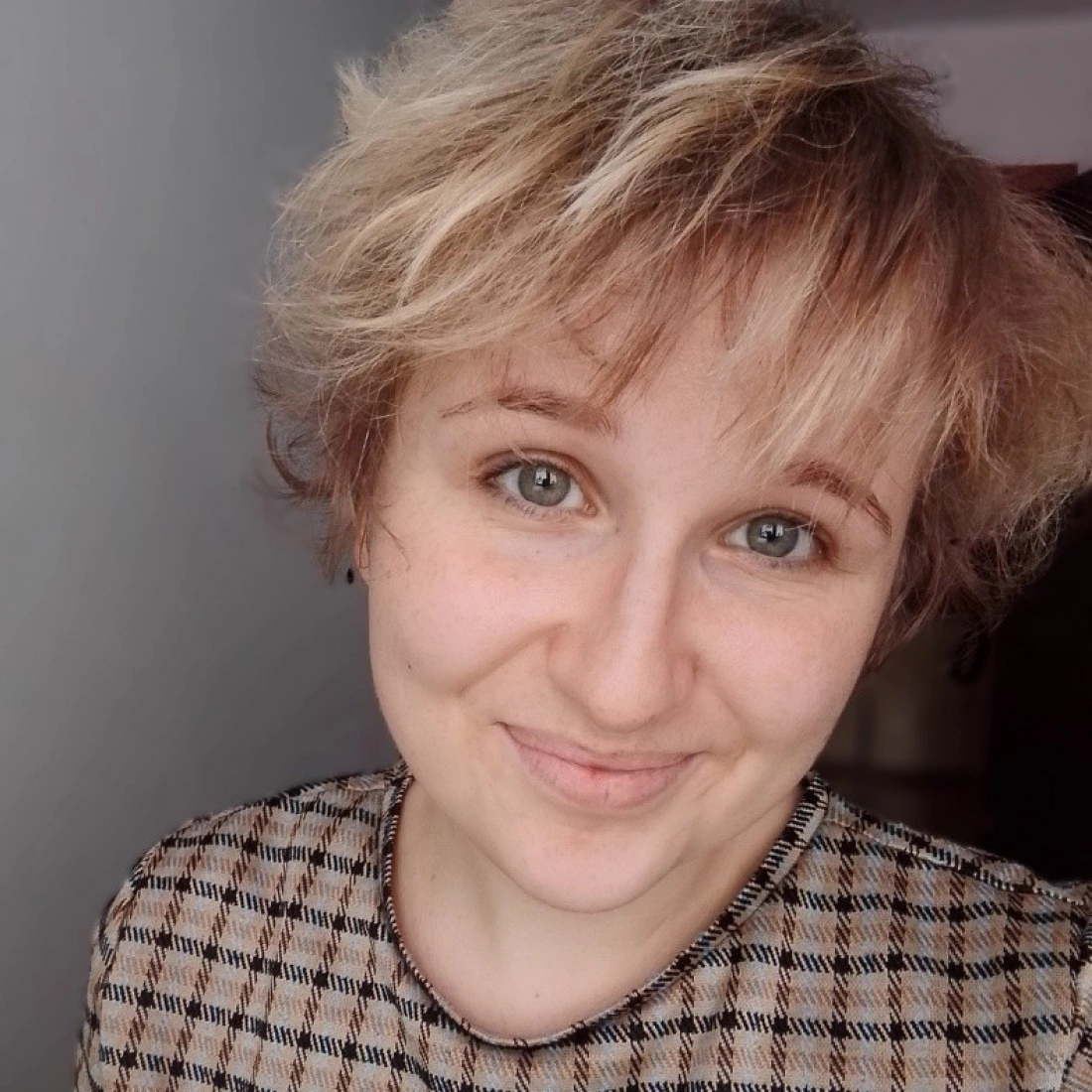 Młoda kobieta z krótkimi, blond włosami uśmiecha się, nosząc wzorzystą bluzkę.