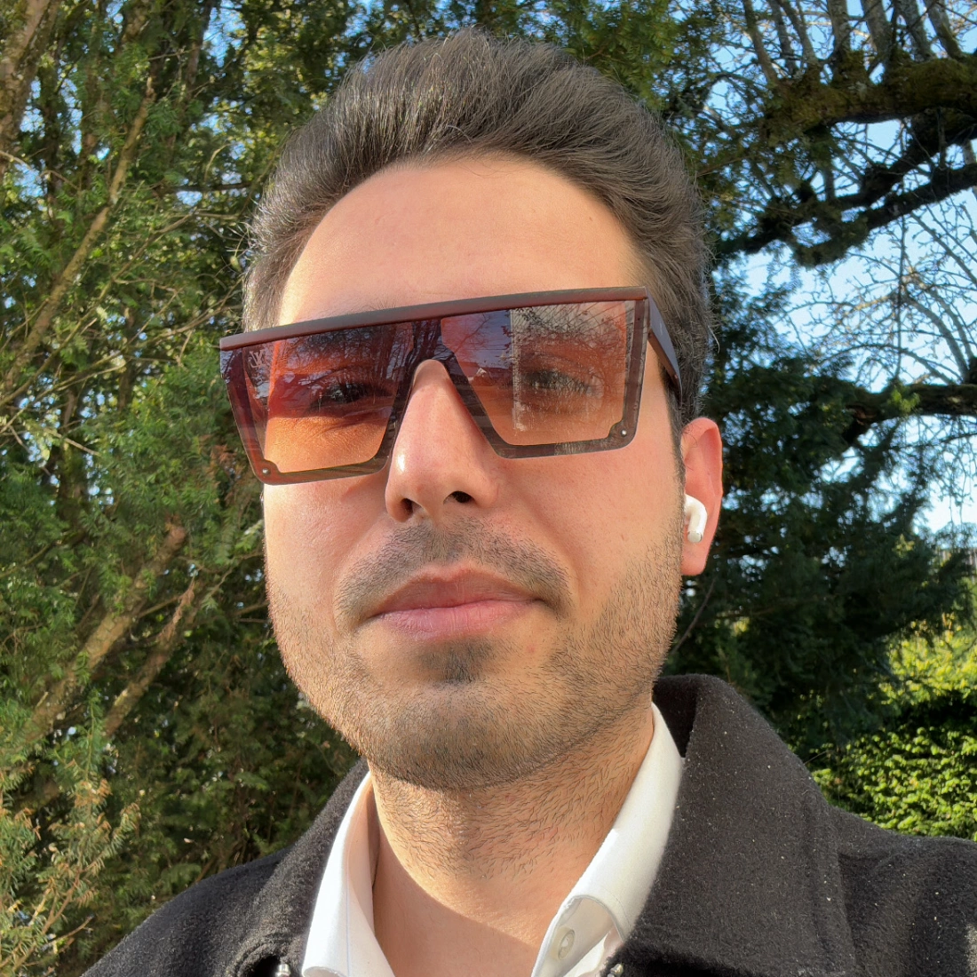 Mężczyzna w dużych okularach przeciwsłonecznych, noszący słuchawki bezprzewodowe, stoi przed zielonym tłem.