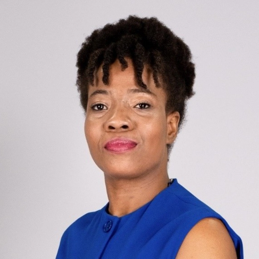 Kobieta o krótkich kręconych włosach, ubrana w niebieską bluzkę, uśmiecha się. Tło jest jasnoszare.