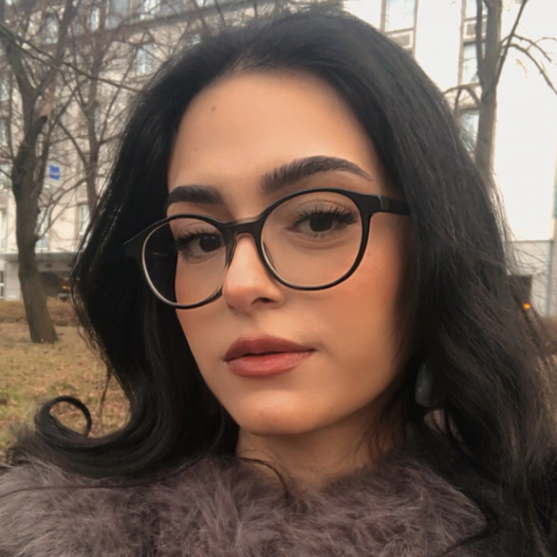 Portret kobiety w okularach, z długimi, ciemnymi włosami, w otoczeniu drzew i budynków.
