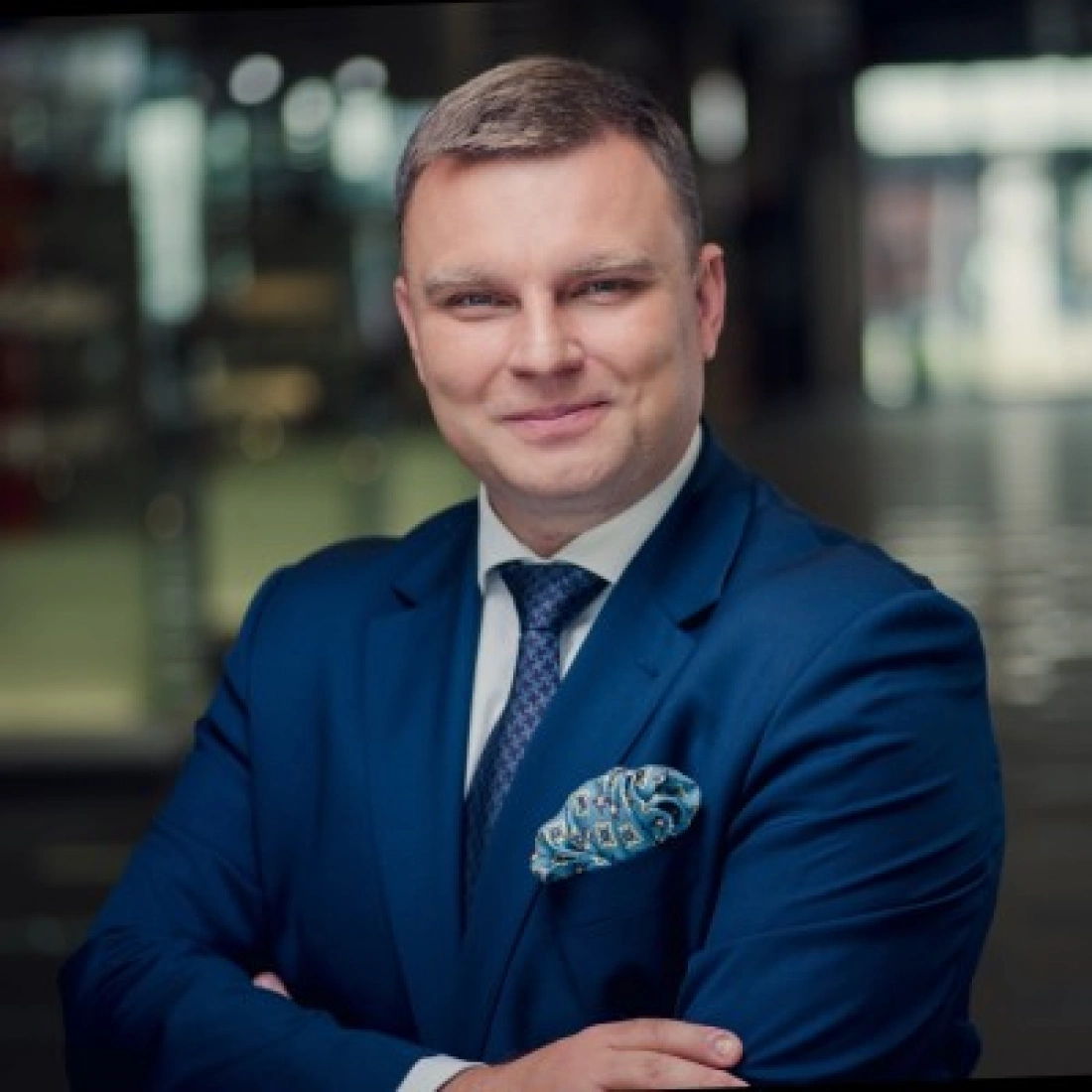 Mężczyzna w niebieskim garniturze z kolorową poszetką, uśmiechający się z założonymi rękami.
