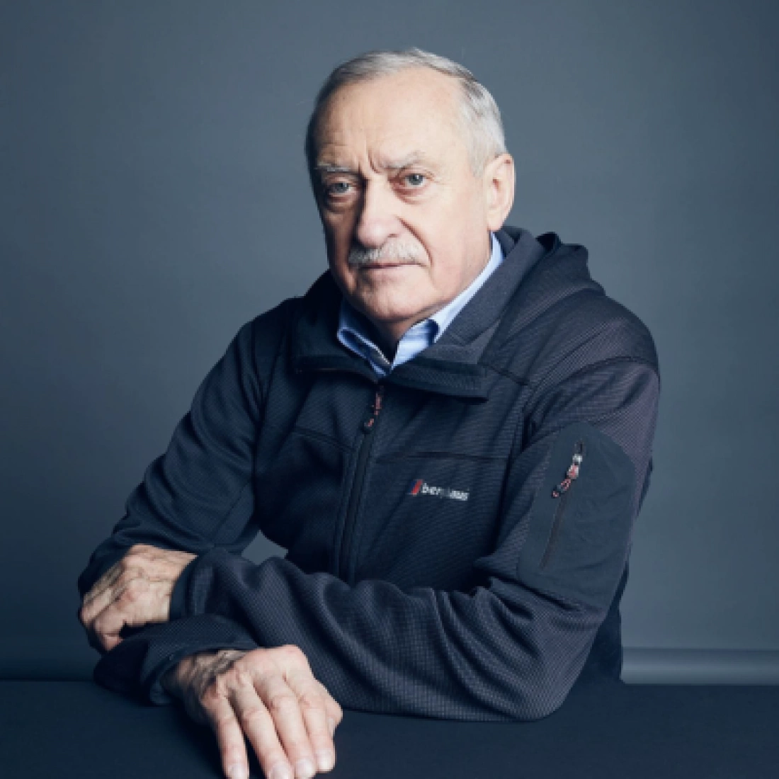 Mężczyzna w czarnej kurtce, siedzący przy stole, z poważnym wyrazem twarzy.