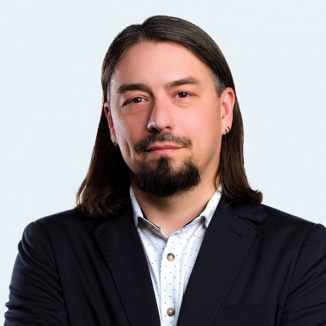 Mężczyzna w garniturze z długimi włosami i zarostem, stojący z założonymi rękami na tle jasnoszarym.