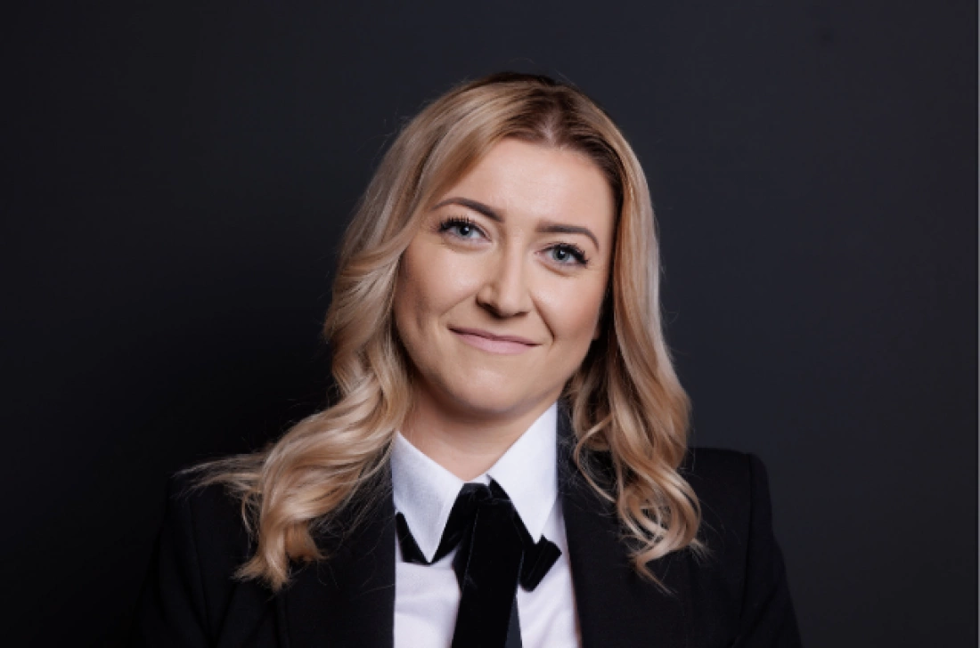 Kobieta w eleganckim stroju, uśmiechająca się na tle ciemnego tła.