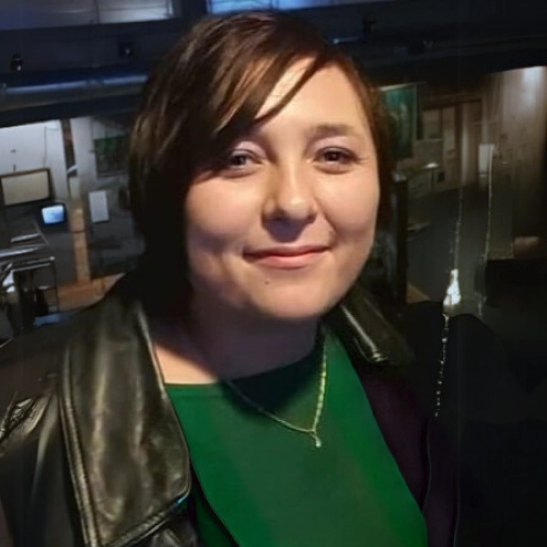 Osoba uśmiechająca się, ubrana w czarną skórzaną kurtkę i zieloną bluzkę, w tle widać galerie sztuki.