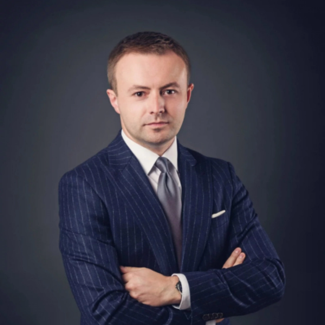 Mężczyzna w garniturze, z rękami skrzyżowanymi na piersiach, z poważnym wyrazem twarzy.