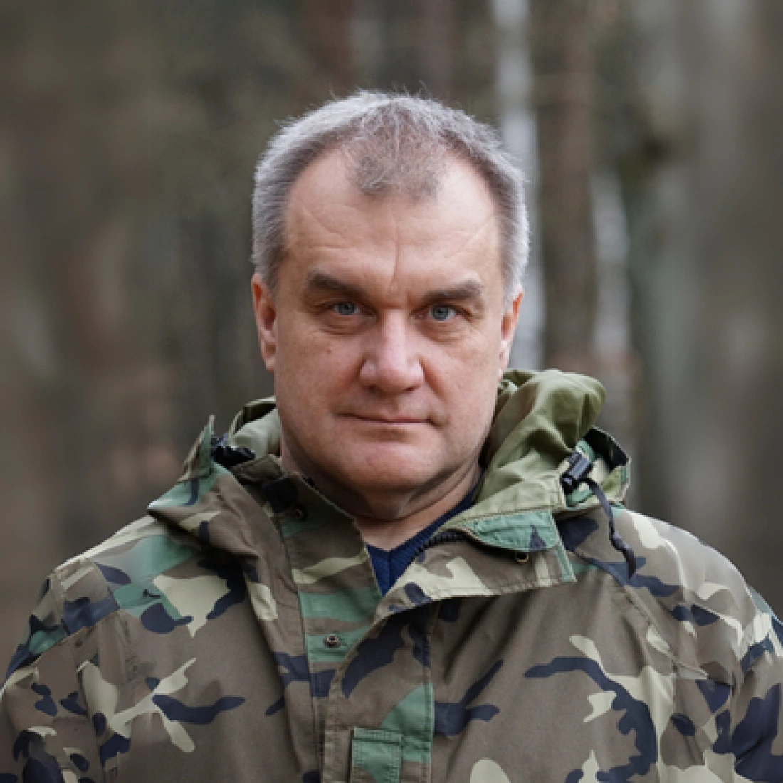 Mężczyzna w mundurze kamuflażowym, stojący w lesie.