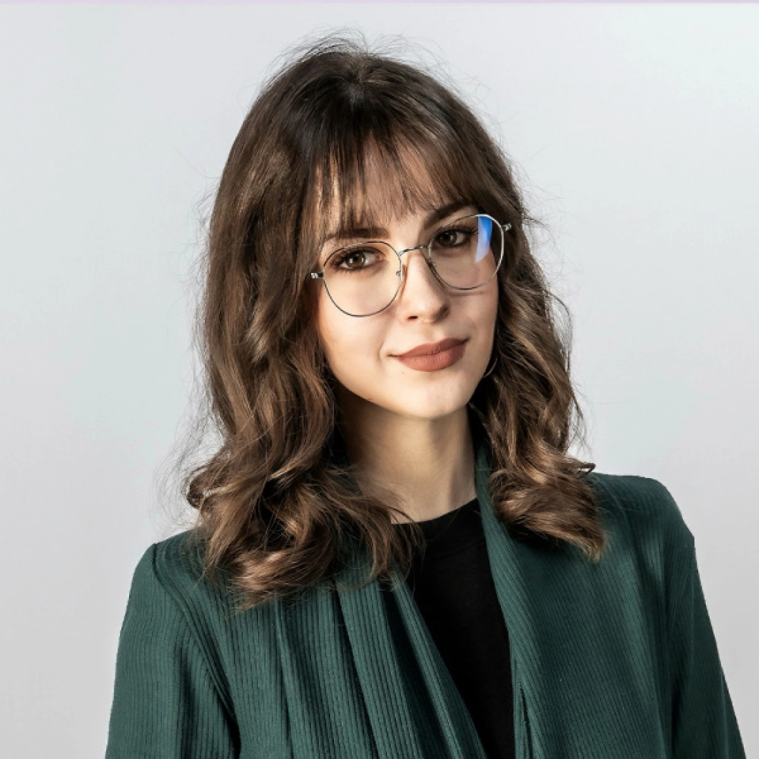 Młoda kobieta z brązowymi falowanymi włosami i okularami, ubrana w zielony żakiet.