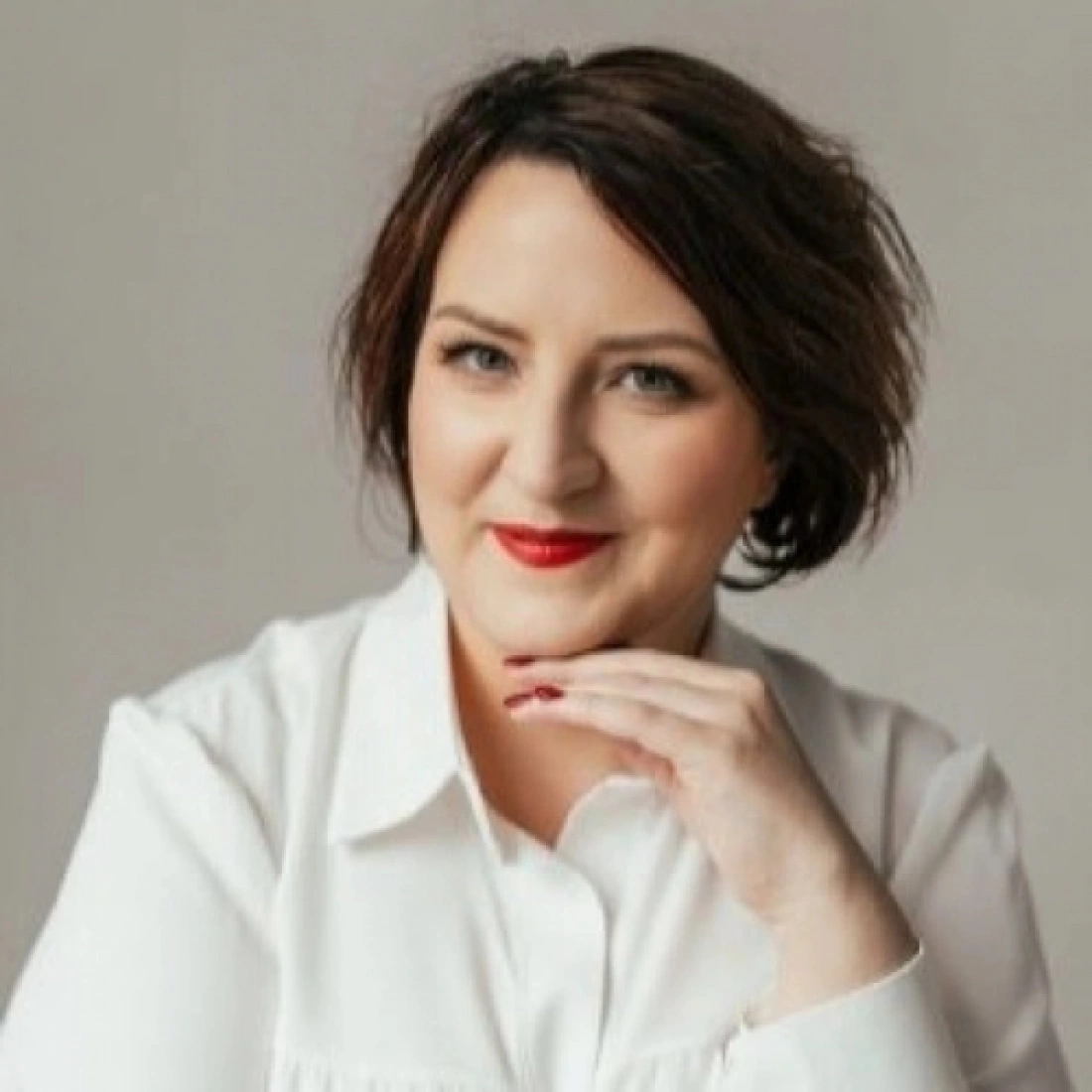 Kobieta o krótkich włosach, uśmiechająca się z ręką na podbródku, ubrana w białą koszulę.