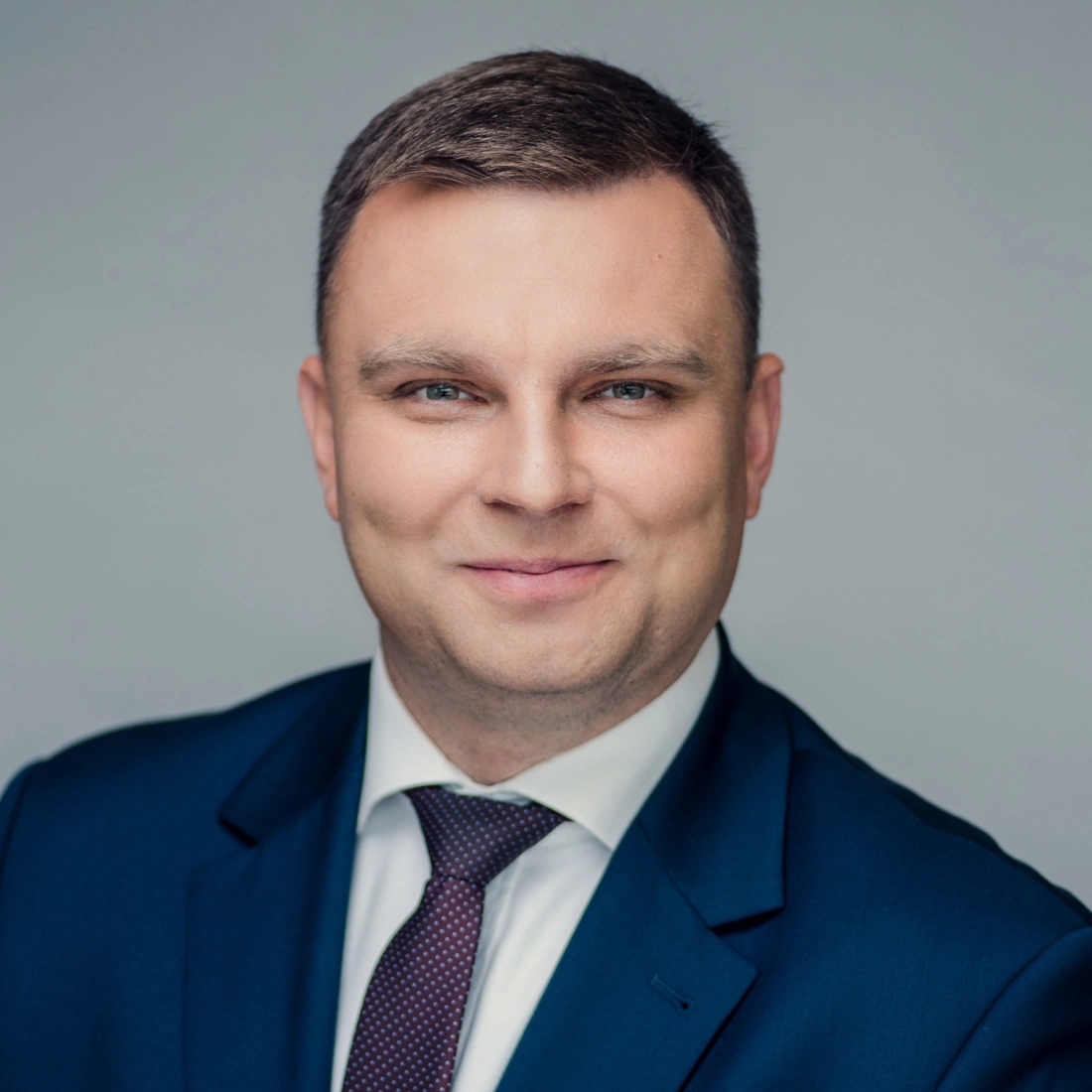 Mężczyzna w garniturze, uśmiechający się do zdjęcia na tle w jasnym kolorze.