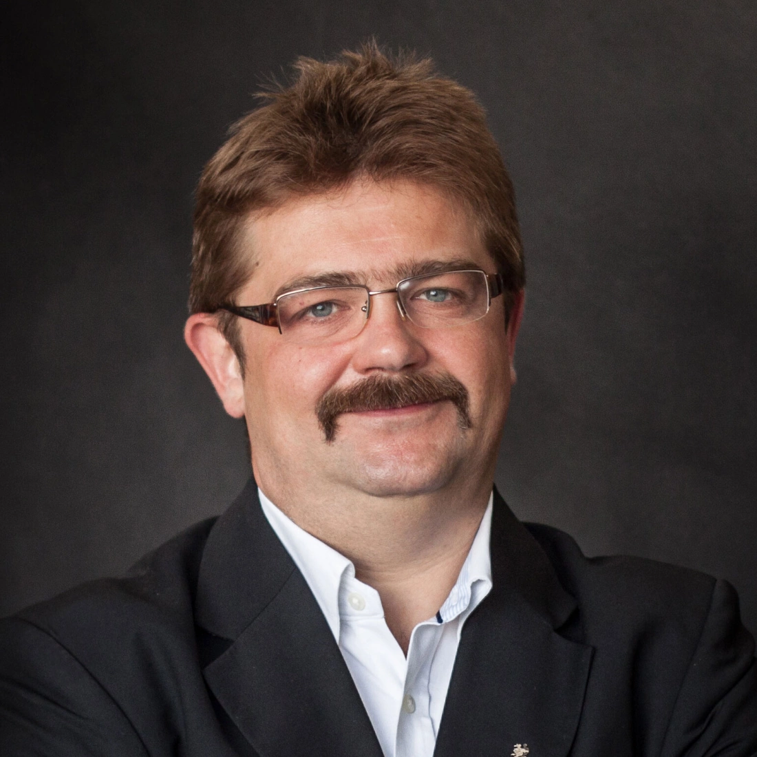 Mężczyzna w okularach, z wąsami, uśmiechający się, ubrany w czarny żakiet i białą koszulę.