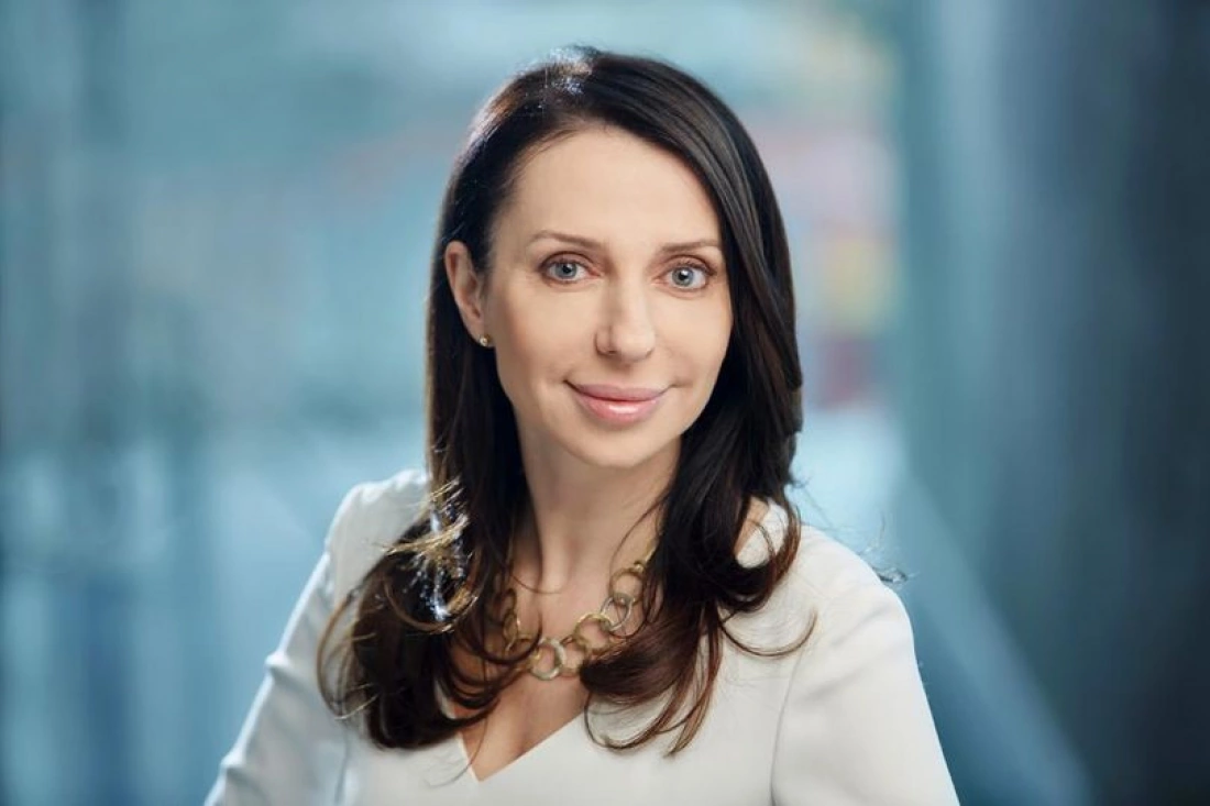 Portret uśmiechniętej kobiety w jasnej bluzce, o długich ciemnych włosach, na tle rozmytej przestrzeni.