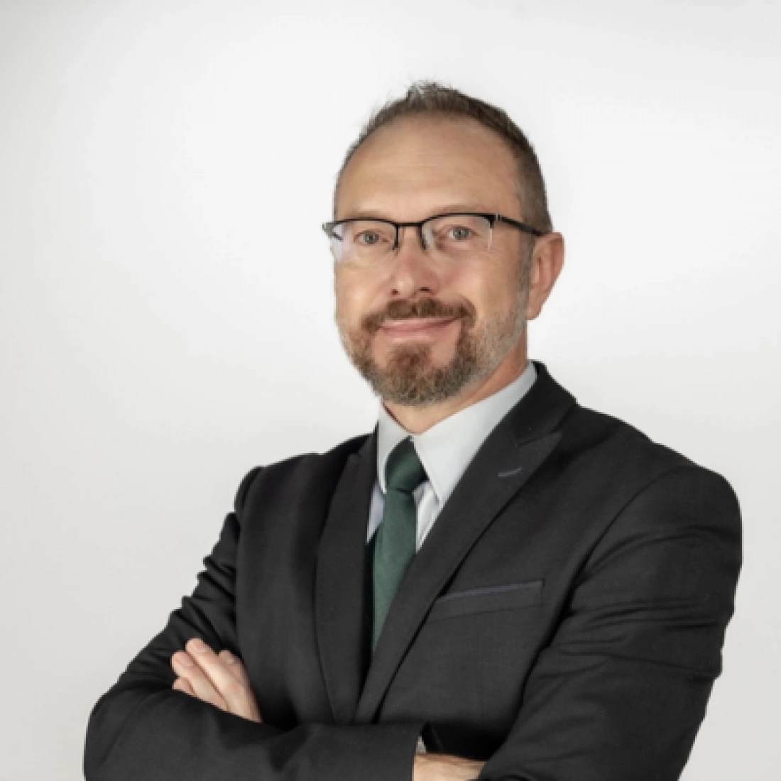 Mężczyzna w garniturze z okularami, stojący z założonymi rękami, na tle jasnej ściany.